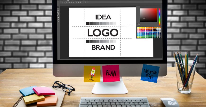Postazione di lavoro con monitor che mostra layout di un logo, ideazione di un payoff, block notes e matite colorate.